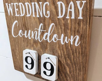 May include: A square wooden sign with a dark brown stained finish featuring the white text WEDDING DAY Countdown. Two small white rectangular wooden tiles with the black number 9 are attached to the front with metal pins to display the count.