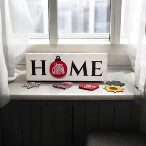 May include: A white wooden sign with the word "HOME" in black letters. A red ornament with "Merry Christmas" text is attached to the "O". Decorative wooden shapes, including a snowflake, heart, and sunflower, are arranged below.
