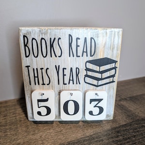 May include: A white wooden sign with the text "BOOKS READ THIS YEAR" and a black silhouette of stacked books. Three white wooden number tiles with black numbers "503" are attached to the sign.