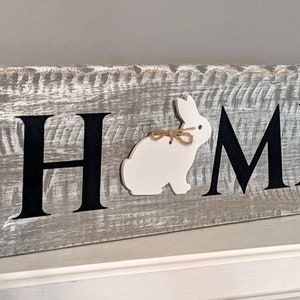 May include: A rustic wooden sign with the word "HOME" in black letters. A white bunny with a twine bow is placed between the "H" and "M". The sign is painted gray with white distressing, creating a farmhouse aesthetic.