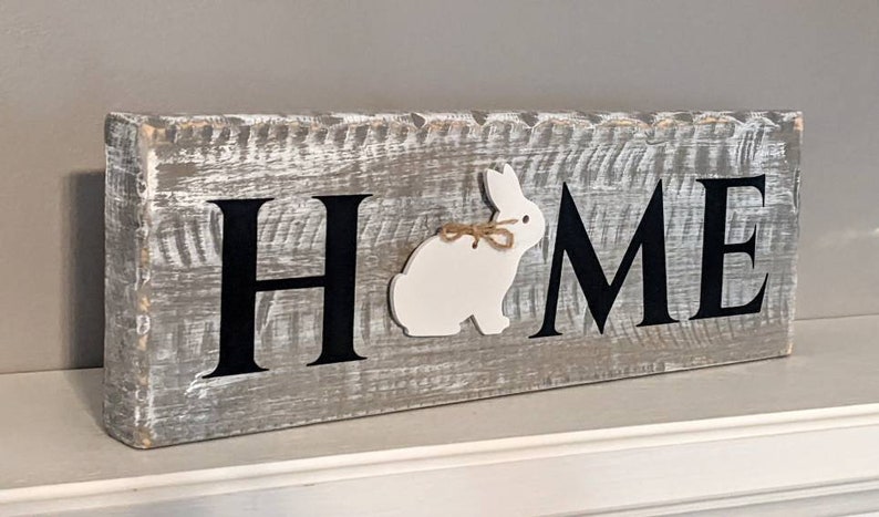 May include: A rustic wooden sign with the word "HOME" in black letters. A white bunny with a twine bow is placed between the "H" and "M". The sign is painted gray with white distressing, creating a farmhouse aesthetic.