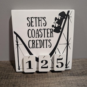 May include: White coaster credit sign with black text that reads "Seth's Coaster Credits". A black roller coaster graphic is on the right. Below are three small white tags with the numbers 1, 2, and 5.