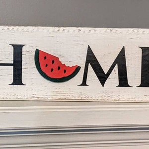 May include: A rectangular wooden sign with the word "HOME" in black letters. The "O" is replaced by a watermelon slice. The sign has a distressed, white-painted background, suggesting a rustic or farmhouse style.