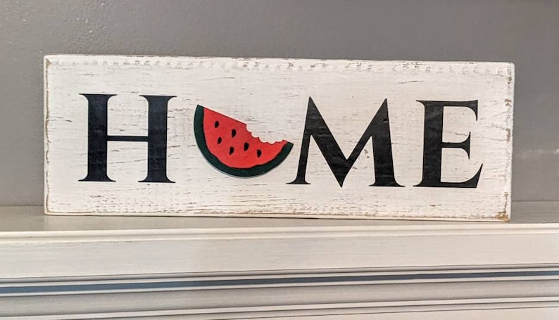 May include: A rectangular wooden sign with the word "HOME" in black letters. The "O" is replaced by a watermelon slice. The sign has a distressed, white-painted background, suggesting a rustic or farmhouse style.