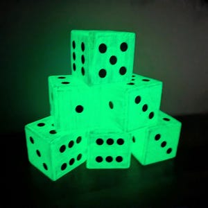 Puede incluir: Una pila de seis dados de madera que brillan con una luz verde brillante en la oscuridad. Cada cubo presenta puntos circulares negros dispuestos en patrones numéricos estándar en sus caras. La textura de la veta de la madera es visible bajo el acabado verde.