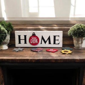 May include: A white wooden sign with the word "HOME" in black letters. A red ornament with "Merry Christmas" is attached to the "O". Decorative wooden shapes, including a snowflake, heart, and sunflower, are arranged below.