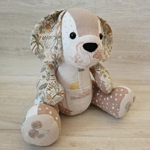 May include: A soft toy dog with a patchwork design. The dog has large, patterned ears, a tan and white face, and a black nose. The body features a mix of floral and polka dot fabrics. The toy is sitting on a light wood surface.