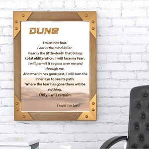 May include: A framed print featuring the word "DUNE" and a quote by Frank Herbert, displayed above a desk with a black leather chair. The quote is set against a desert landscape. A desk lamp and office supplies are also visible.