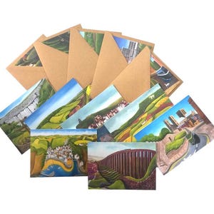 May include: A set of landscape art greeting cards with brown envelopes. The cards display colourful paintings of diverse outdoor scenes, including rolling hills, buildings, and bridges. The envelopes are made from natural, textured paper.
