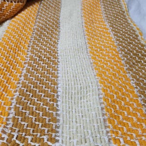 Puede incluir: Un tejido con rayas verticales en tonos amarillo brillante, marrón dorado y crema. La tela muestra un patrón texturizado distintivo creado por la alternancia de hebras de hilo gruesas y finos hilos blancos entrelazados en cruz.