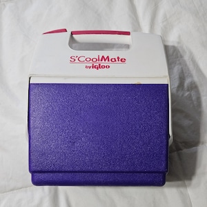 May include: A portable S'CoolMate by Igloo cooler featuring a white top section with a pink handle and a purple textured base. The front of the white lid displays the text S'CoolMate by igloo in pink lettering, providing a compact storage solution.