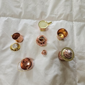 May include: A collection of miniature copper and brass kitchenware items, including small pots, a lidded container, a handled bucket, and a shaker-style piece, all arranged in a scattered formation on a white fabric surface.