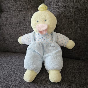 May include: A plush duck toy with a pale yellow head, a small tuft of yellow yarn on top, and blue embroidered eyes. It wears a light blue textured romper with white sleeves featuring blue polka dots and a small blue ribbon at the neckline.
