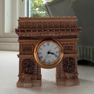 May include: A decorative clock shaped like the Arc de Triomphe. The clock has a gold-colored face with Roman numerals and black hands. The arch is beige and tan with detailed carvings. A vase and flowers are in the background.