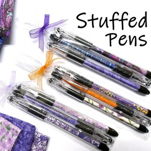 May include: Three sets of pens wrapped in clear plastic with purple and orange ribbon bows. The pens are wrapped in colorful fabric scraps. The text "Stuffed Pens" is written in black at the top of the image. The text "Ami Simms Quilt Studio" is written in white on a red house icon in the bottom right corner of the image.