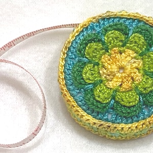 May include: A crocheted measuring tape with a yellow, green, and blue flower design. The tape is white with red markings.