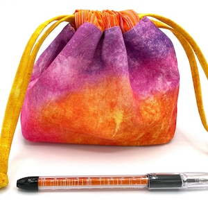 May include: A small drawstring bag made of tie-dye fabric in shades of purple, orange, and yellow. The bag has a yellow drawstring and is sitting on a white surface. A black pen is also pictured.
