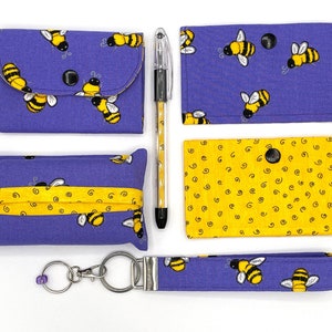 May include: A set of purple and yellow bee-themed fabric items, including a small zippered pouch, a tissue holder, a small wallet, a key chain, and a pen. The items are decorated with bees and a swirl pattern.