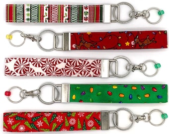 May include: Five different Christmas themed keychains with silver key rings. The keychains are made of fabric with various patterns including candy canes, Christmas lights, and Christmas trees.