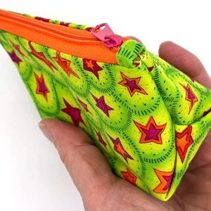 May include: A small, handmade pouch with a vibrant star pattern. The pouch features a lime green background with repeating stars in shades of pink, orange, and red. It has an orange zipper and a pink zipper pull. The pouch is held in a hand.