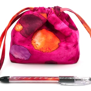 May include: A pink drawstring bag with orange and purple watercolor-like spots. The bag has a long orange drawstring. A black and clear pen is lying in front of the bag.