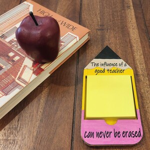 May include: A wooden pencil-shaped notepad holder with a yellow and pink design. The notepad has a yellow sticky note and the text "The influence of a good teacher can never be erased".