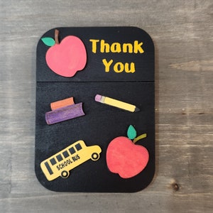 May include: Black wooden sign with the words "Thank You" in yellow letters. The sign is decorated with red apples, a yellow school bus, a purple book, and a yellow pencil.