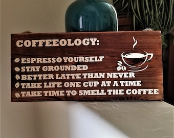 May include: A wooden sign with the text "COFFEEOLOGY:" and a list of coffee-themed puns, including "Espresso Yourself", "Stay Grounded", "Better Latte Than Never", "Take Life One Cup At A Time", and "Take Time To Smell The Coffee".