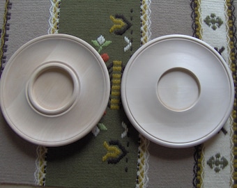 May include: Two unfinished, light-colored wooden plates. One plate has a raised center with a circular indentation, while the other has a simple circular hole in the center. The plates are on a patterned fabric.