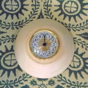 May include: A round wall clock featuring a smooth, light-colored wooden frame and a gold-toned bezel. The clock face has a white background with black Arabic numerals, a central gold-toned circular accent, and black hour, minute, and second hands.