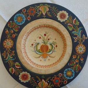 May include: A round wooden plate with a dark blue rim and a cream-colored center. The plate is decorated with hand-painted floral designs in shades of red, orange, yellow, green, and blue.