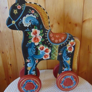 May include: A hand-painted wooden toy horse with four wheels. The horse is black with blue, red, and yellow floral designs. The wheels are red with blue and gold accents.