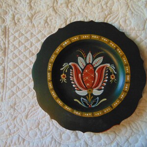 May include: A black wooden plate with a scalloped edge, painted with a stylized red, white, and green flower design.