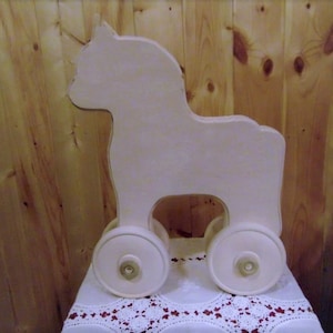 May include: A white wooden toy horse with four wheels. The horse is standing on a white lace tablecloth.