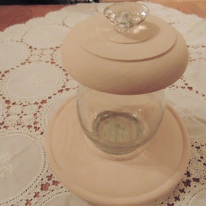 May include: A white ceramic jar with a glass lid and a knob on top. The jar is sitting on a white lace tablecloth.