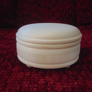 May include: A small, round, wooden box with a lid. The box is made of light-colored wood and has a smooth finish. It has three small feet on the bottom.
