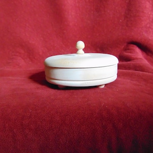 May include: A small, round, light-colored wooden box with a lid and a small knob on top. The box sits on three small feet. The background is a solid, deep red color.