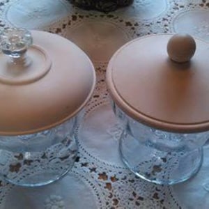 May include: Two glass candy dishes with lids. One lid has a clear, decorative knob, and the other has a small, round wooden knob. The lids are a light beige color, and the dishes sit on a white lace tablecloth.