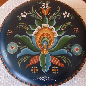 May include: A round wooden plate with a hand-painted design. The plate is painted in shades of blue, green, orange, and white. The design features a stylized flower with leaves and a border of dots.