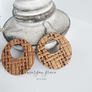 May include: A pair of tan-colored, woven-patterned hoop earrings. The earrings have a circular design with a cutout in the center and silver-colored ear wires. The text "morgan grace DESIGNS" is printed on the surface.