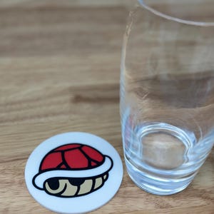May include: A white, round coaster featuring a red turtle shell design with a black outline and tan accents. A clear glass sits to the right on a wooden surface. The coaster is a fun accessory for any beverage.