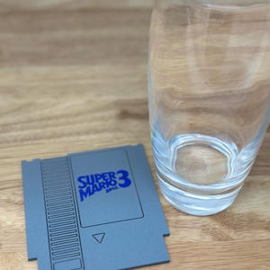 May include: A gray and black Super Mario Bros. 3 video game cartridge coaster. The coaster is shaped like a NES game cartridge and has the Super Mario Bros. 3 logo printed on it in blue.