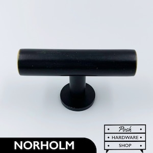 May include: A black cabinet pull with a cylindrical handle and a circular base. The handle has a brass-colored end. The word "NORHOLM" is printed in white on a black rectangle. The image also includes the "Posh HARDWARE SHOP" logo.
