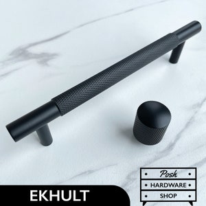 Peut inclure: Poignée de meuble noire avec une finition texturée. La poignée mesure 15 cm de long et a un bouton rond à l'extrémité. Le texte "EKHULT" est visible sur l'image.