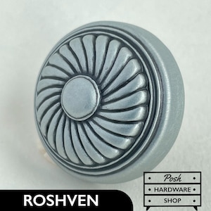 Könnte beinhalten: Ein runder Schrankknopf in Silberton mit einem dekorativen, erhabenen Sonnenstrahl-Design. Der Knopf hat eine glatte, leicht strukturierte Oberfläche.