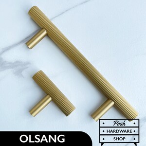 May include: Two gold cabinet pulls with a ribbed design. The larger pull is 12.7 cm long and the smaller pull is 7.6 cm long. The pulls are on a white marble background. The text "OL SANG" is in the bottom left corner of the image. The text "Posh Hardware Shop" is in the bottom right corner of the image.