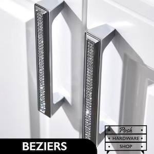 May include: Two silver cabinet door pulls with a row of clear crystals down the center. The pulls are mounted on a white cabinet door. The text "BEZIERS" is visible at the bottom of the image.