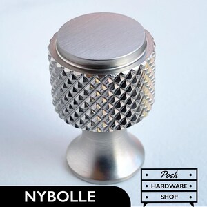 Op de afbeelding: Een geborstelde zilveren kastknop met een getextureerde, diamantvormige grip. De knop heeft een platte, cirkelvormige bovenkant en een afgeronde basis. De tekst "NYBOLLE" wordt weergegeven in een zwarte rechthoek aan de onderkant van de afbeelding. Het logo "Posh HARDWARE SHOP" is ook zichtbaar.
