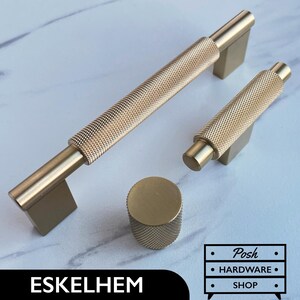 Op de afbeelding: Drie goudkleurige kastbeslagstukken: een grote en een kleine greep met een getextureerd, geribbeld ontwerp, en een ronde knop met een vergelijkbaar getextureerd ontwerp. De tekst "ESKELHEM" is zichtbaar in de afbeelding.