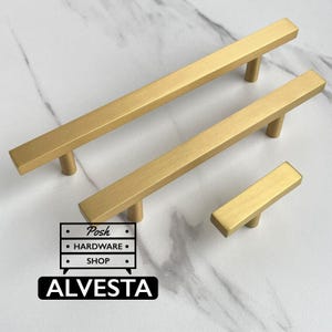 May include: Three brushed gold cabinet pulls of varying lengths, each featuring a linear, rectangular bar design with two cylindrical mounting posts. A logo graphic is present, displaying the text Posh HARDWARE SHOP and ALVESTA.